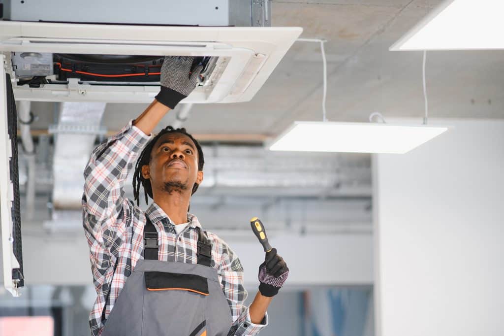 African american hvac technician is working on an air conditioning unit, performing maintenance and ensuring proper ventilation in a modern office environment