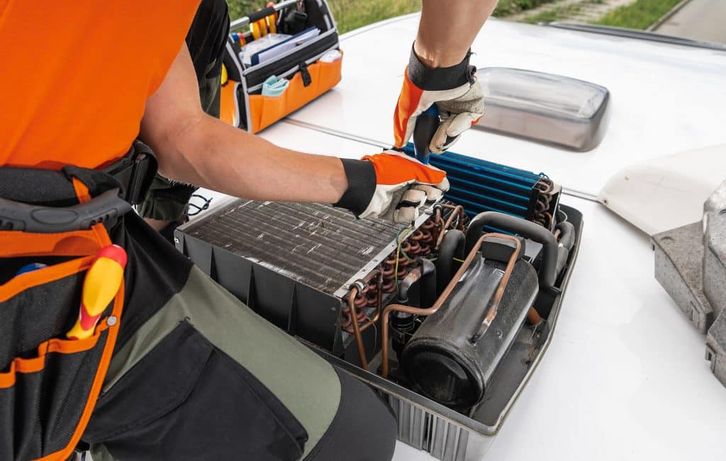 A technician adjusts components of a rooftop RV Camper HVAC unit, using tools from an open toolbox, while working on a building.