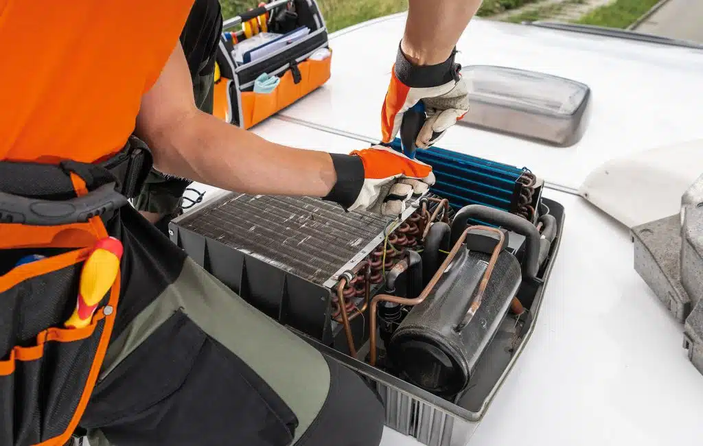 A technician adjusts components of a rooftop RV Camper HVAC unit, using tools from an open toolbox, while working on a building.
