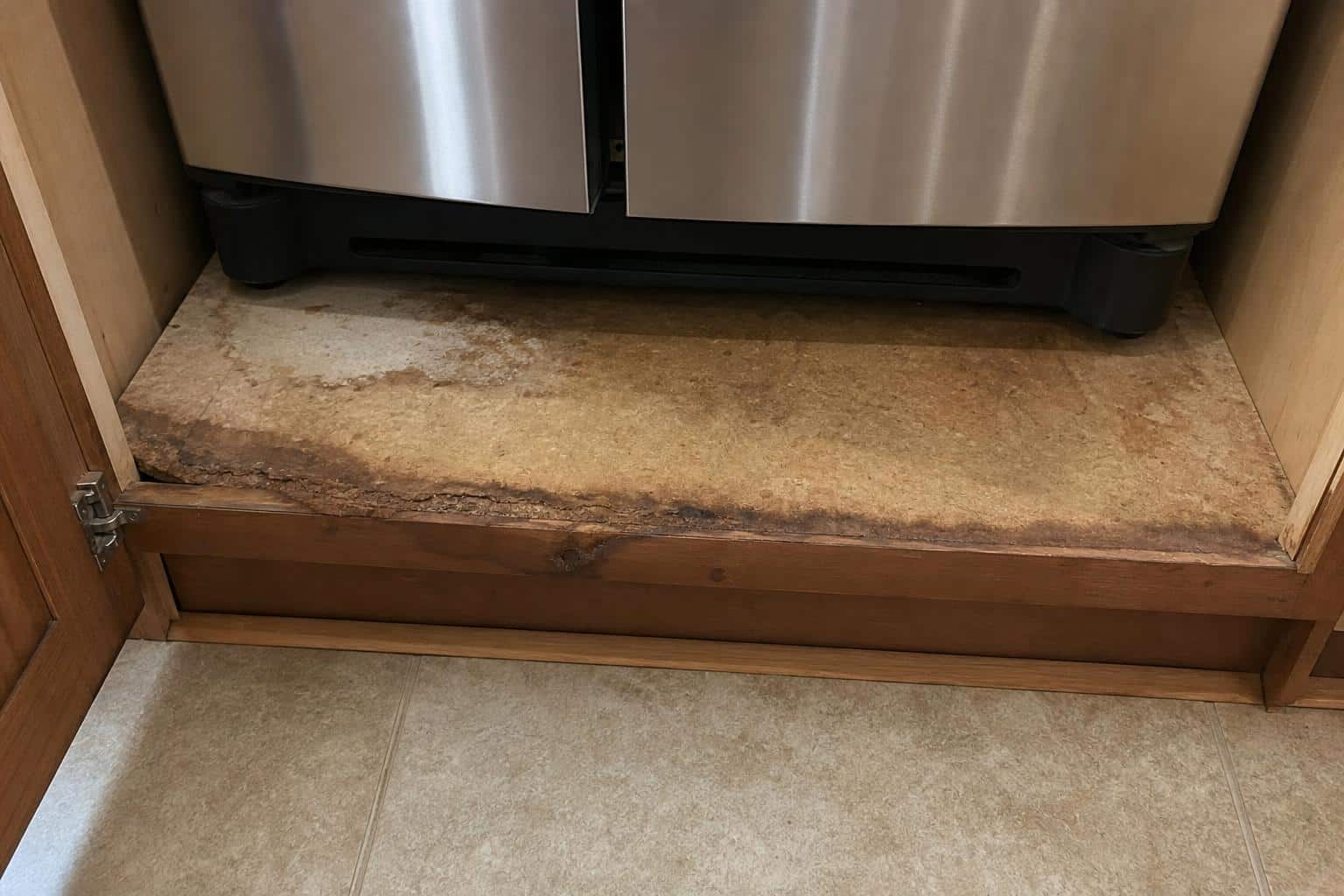Water-stained particleboard cabinet base beneath a kitchen refrigerator showing swelling and discoloration
