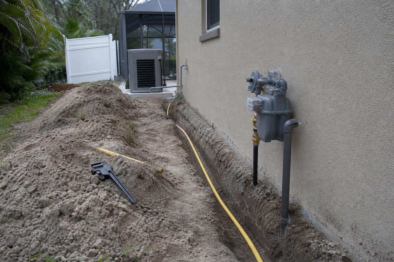 A trench alongside a Florida home's exterior wall with black gas supply pipe being laid toward a generator pad, daytime natural light, no readable text, no brand logos, no people's faces