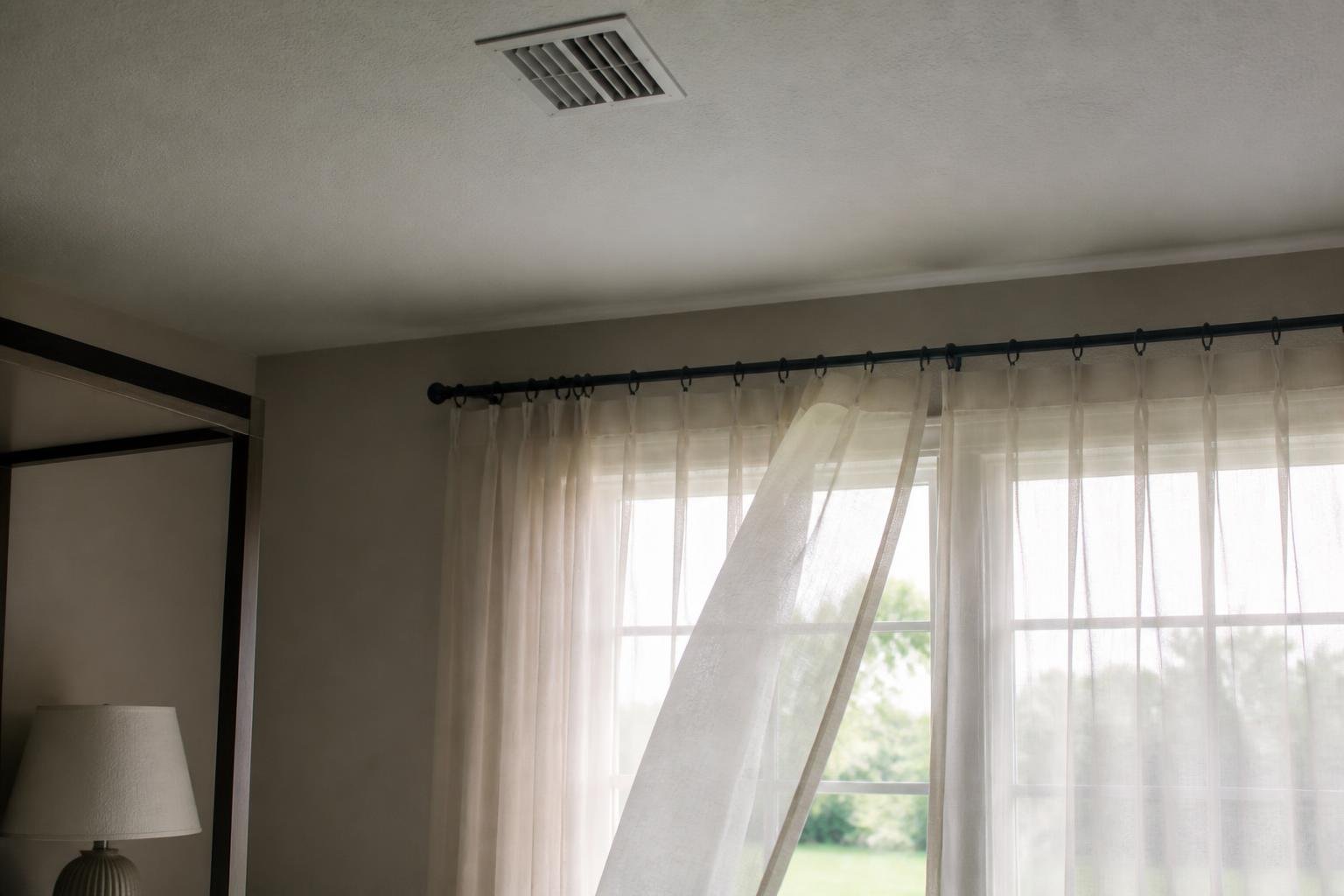 A ceiling air vent register in a Florida bedroom with visible airflow movement suggested by a light curtain nearby, soft daylight, no readable text, no brand logos, no people's faces